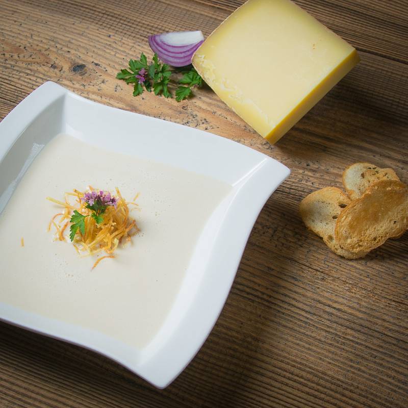 Bergkäsesuppe in einem Teller, daneben Ein Stück Käse und Brotchips